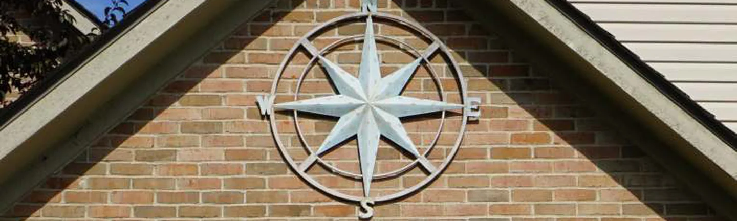 The front gable of the Michigan house in 2019, a metal compass rose mounted on tan brick that reads as one range under the roofline