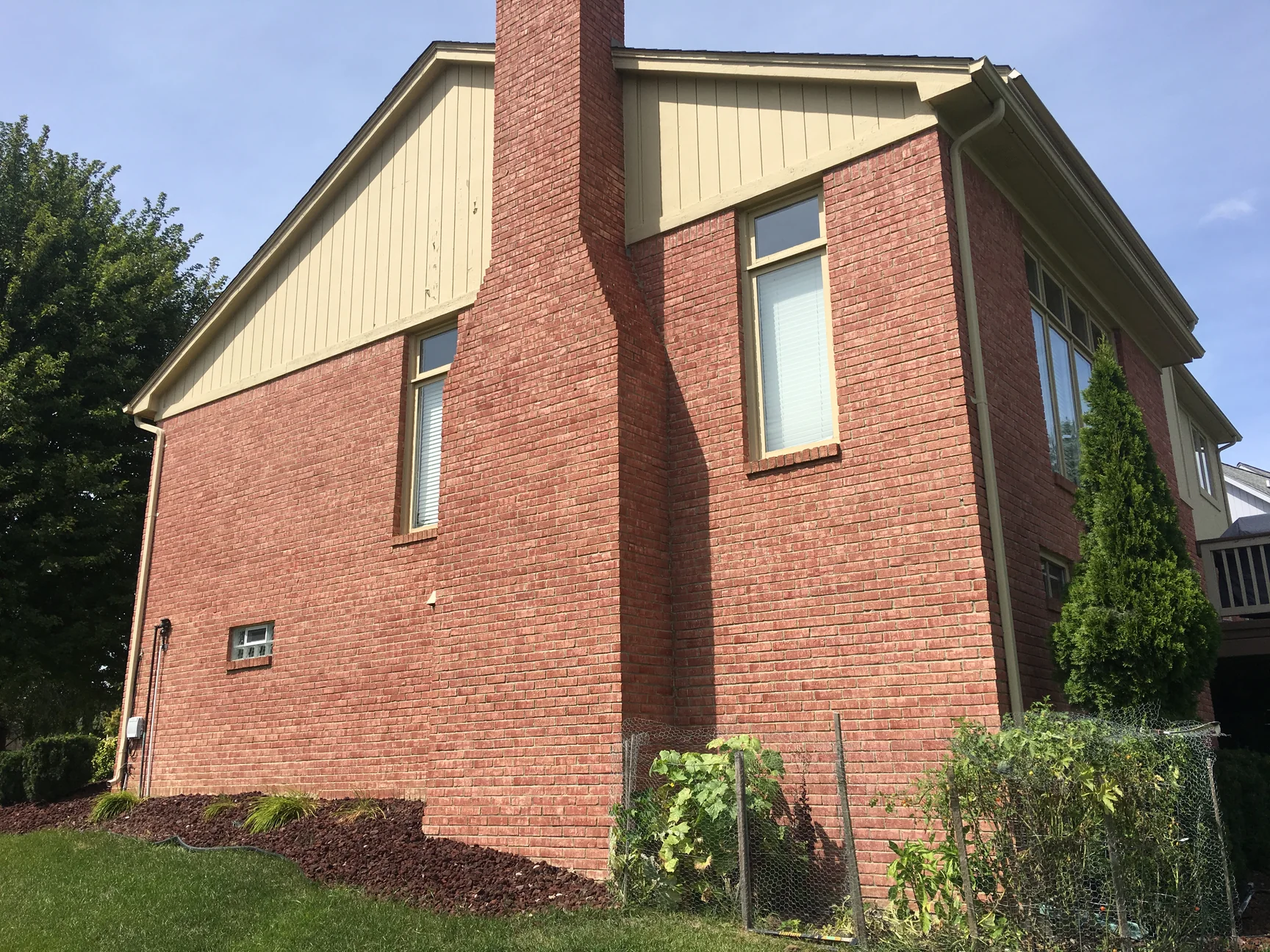The chimney side in 2019, salmon brick across the full wall and up the chimney to the cream gable siding, a small basement window low on the wall and a garden fence at the right