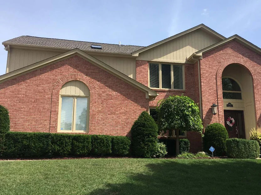 The front of the two-story house in 2019 from the lawn, salmon brick under cream siding, an arched entrance bay at the right and a clipped hedge along the foundation