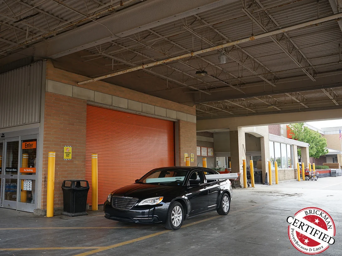The covered entry bay in 2019, the brick pier beside the orange rolling door holding one red and tan range, the frame carrying the Brickman Certified stamp