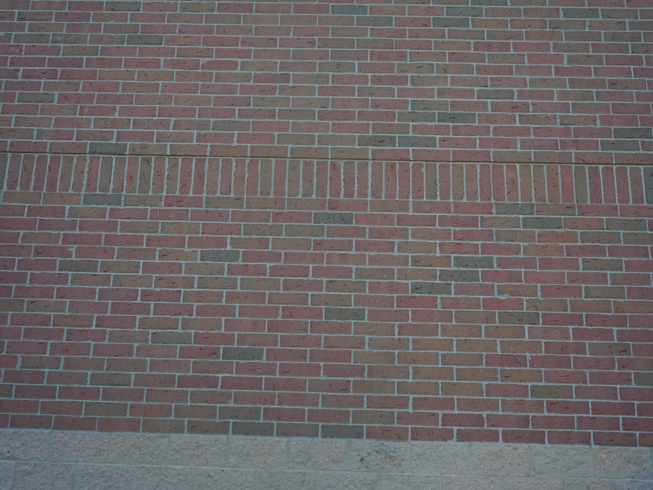 Close view of the wall in 2019, red and grey brick with a course of upright units running through it above a cast stone base, the joints pale and unbroken