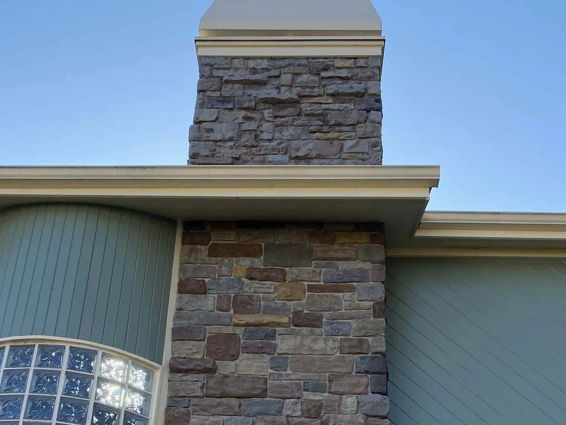 The same chimney in 2021, the stone now a blend of brown, rust, grey and tan, running down past the eaves to the curved glass block bay below