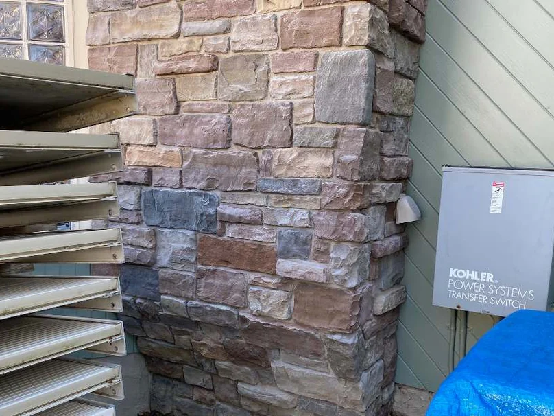 The same side wall in 2021, the stone in a brown, rust and blue grey blend, the Kohler transfer switch and the glass block window still beside it