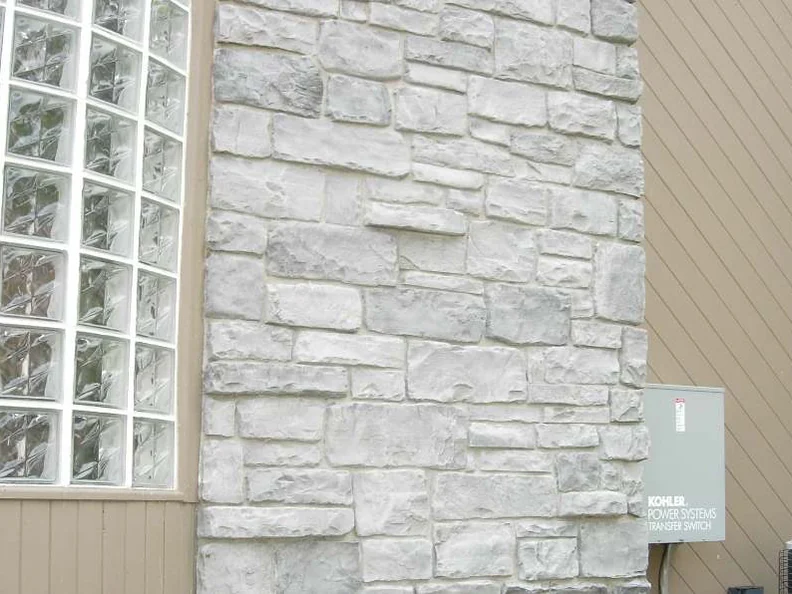 A side wall in 2007, pale grey manufactured stone between a curved glass block window and a Kohler transfer switch mounted on tan siding
