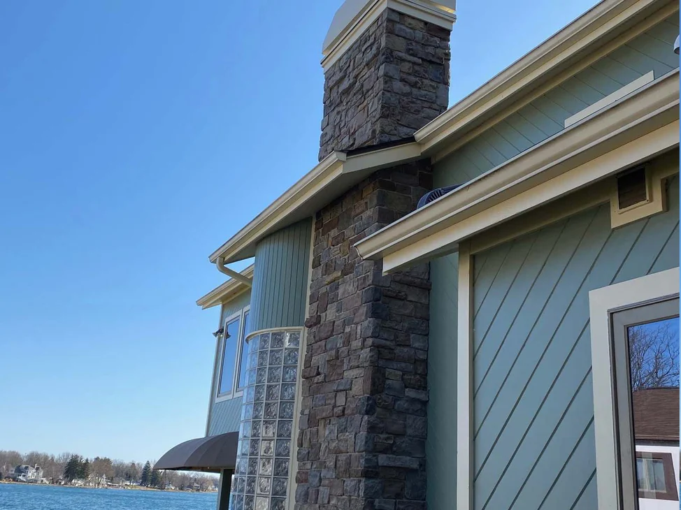 The lake elevation of the Michigan house in 2021, a stone chimney over a full height stone column in a brown, grey and tan blend, green siding either side and open water beyond