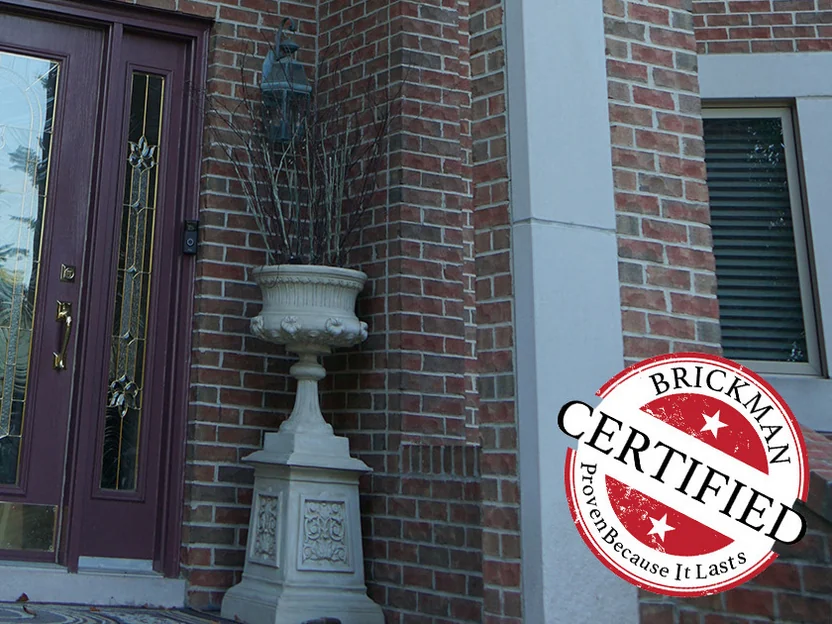 The front door in 2019 with a leaded glass sidelight, a stone urn beside it and the brick reveal and outside corner running past in one red and brown range, the certification stamp printed at the corner of the frame