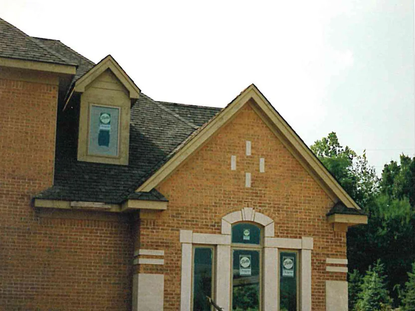 The gable end of the same house in 1996, its brick a warm orange tan, cast stone blocks set in the gable field above an arched window group, with graded soil and a pickup truck at the foot of the wall