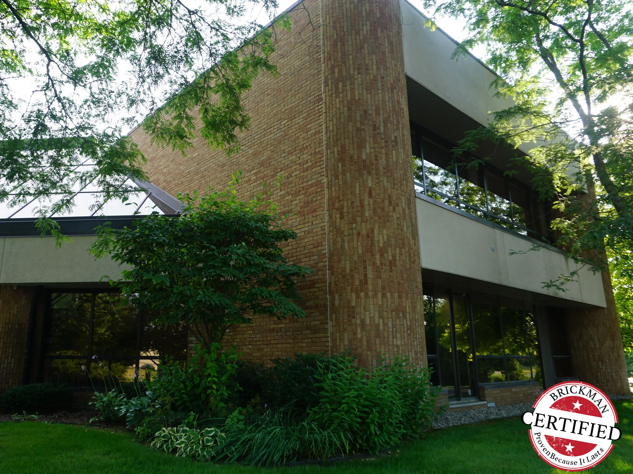 The outside corner of the office under mature trees in 2019, the rounded brick pier and the flat wall it turns in one range, with the certification stamp in the frame