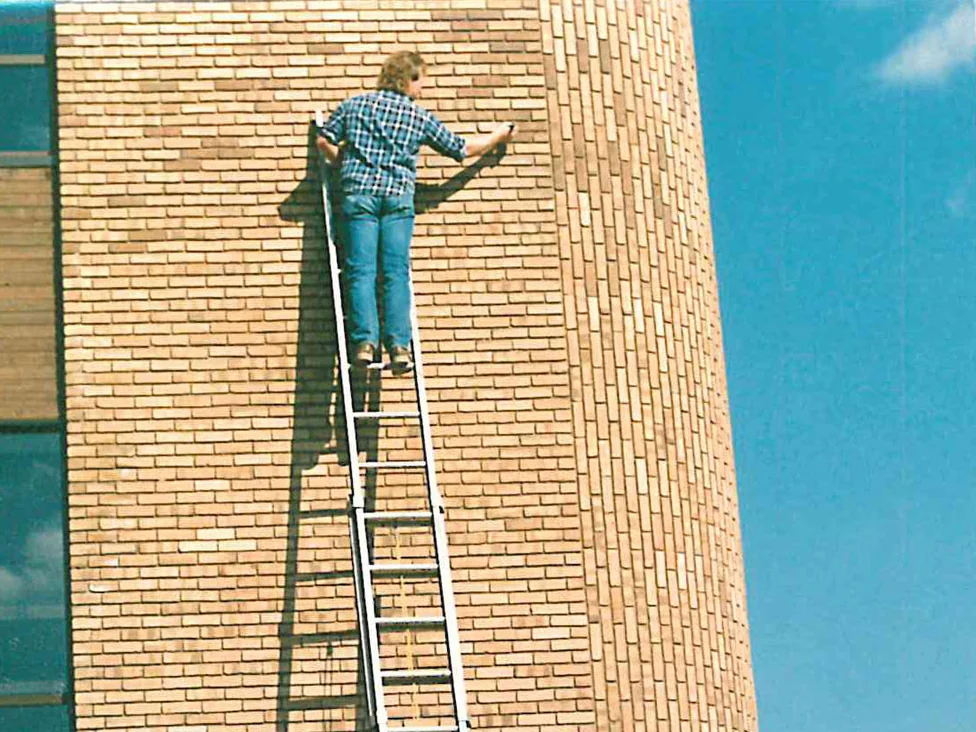 An applicator standing high on a ladder working across the brick beside the rounded corner of the office, the ground around the building still bare