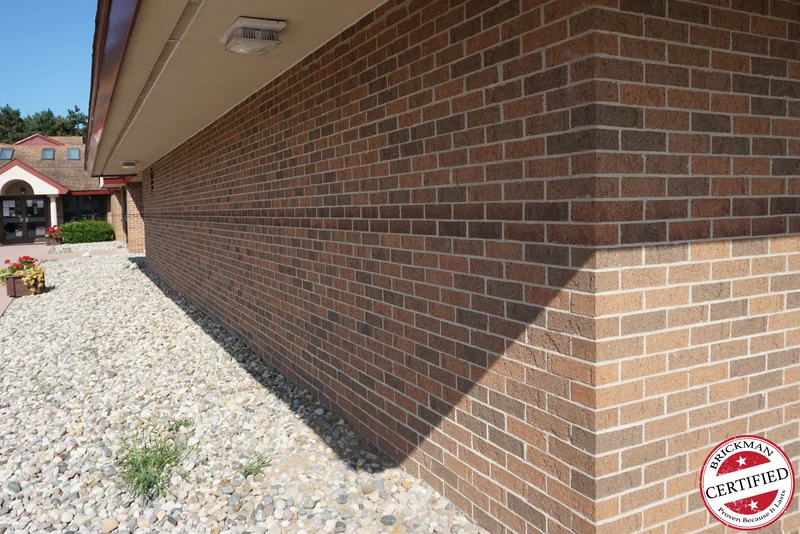 The wall in 2019 running away to an outside corner, a band of darker brick carrying round the corner above lighter buff courses, the entrance canopy in the distance and the certification stamp at the corner of the frame