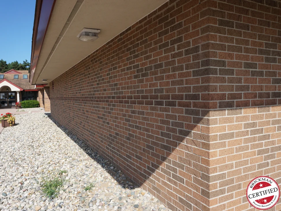 The wall in 2019 running away to an outside corner, a band of darker brick carrying round the corner above lighter buff courses, the entrance canopy in the distance and the certification stamp at the corner of the frame