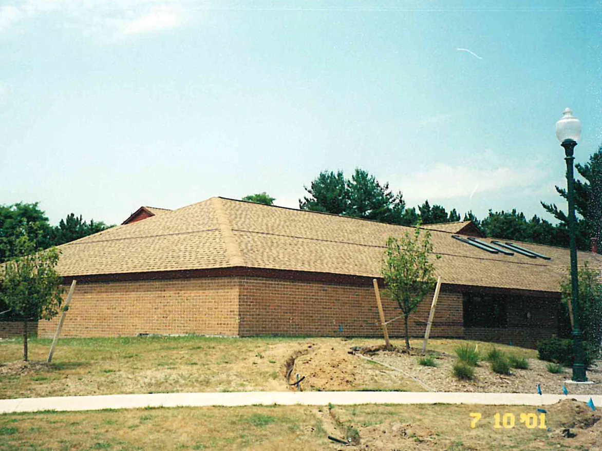 The office in 2001 before the work, one flat buff tan brick running across the end wall and the wing beyond it, the site newly planted with staked trees and bare soil