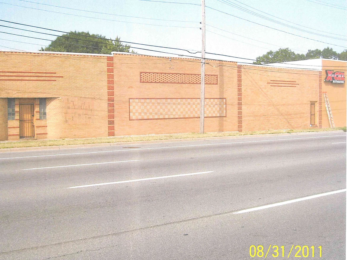 A long single-story brick building seen from across a wide road in 2011, the wall one flat tan range with its decorative panels barely showing