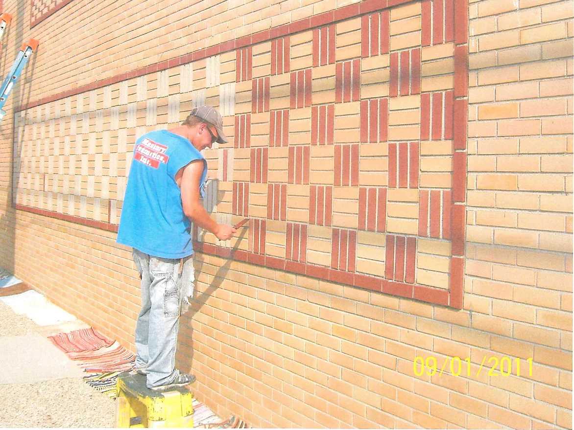 An applicator on a step stool brushing stain onto individual bricks in a basketweave panel, the finished red pattern to his right and the pale untouched coursing to his left
