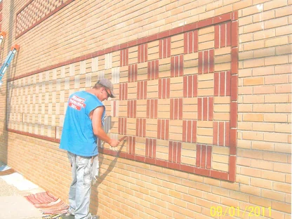 An applicator on a step stool brushing stain onto individual bricks in a basketweave panel, the finished pattern to his right and the pale untouched coursing to his left