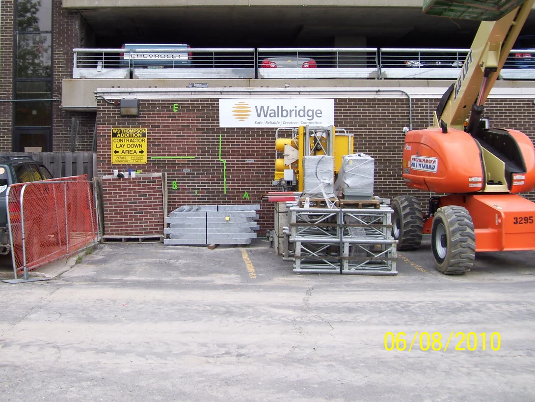 The base of the parking garage in June 2010, dark brown brick under a Walbridge sign with a contractor lay-down area in front, stacked steel, a fenced yard and a lift parked at the wall
