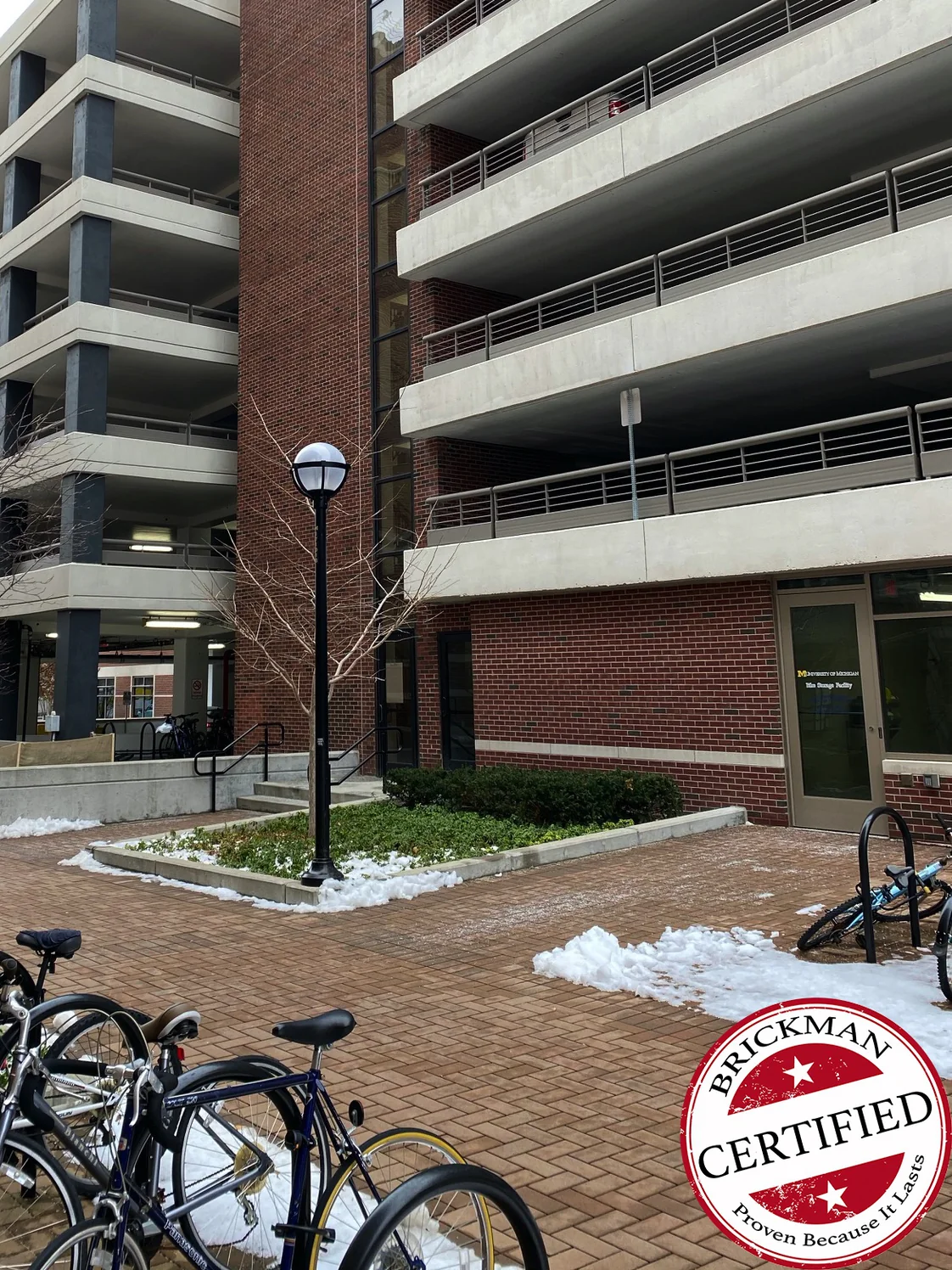 The parking garage tower in 2020, one range of red brick from the upper decks down to the glazed entrance door, bike racks and snow on the paving in front and the certification stamp in the corner