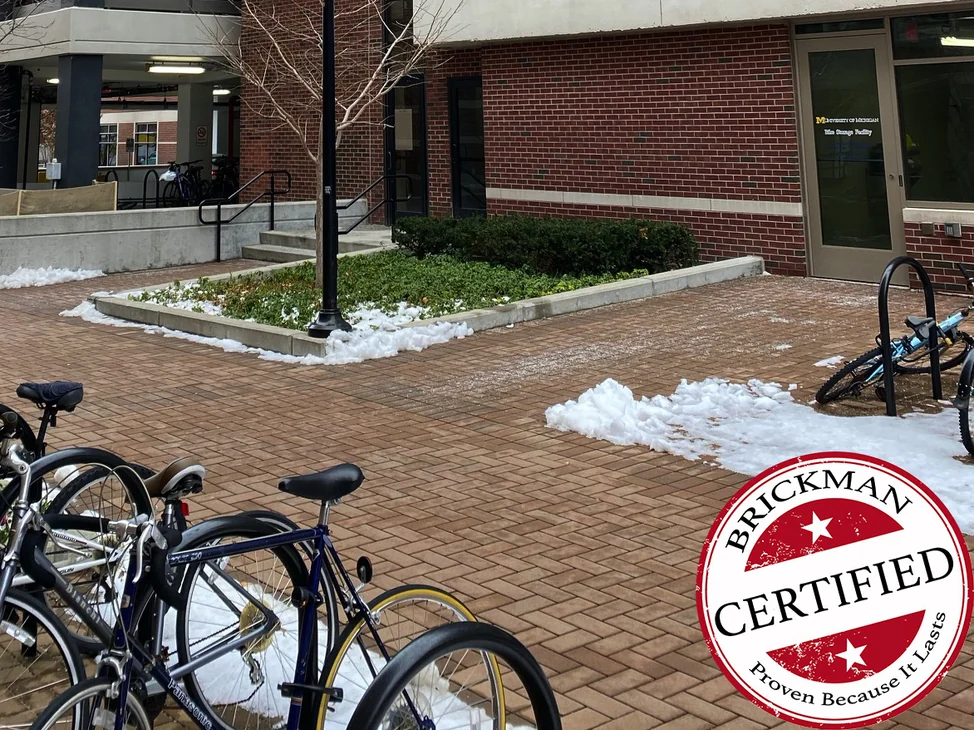 The base of the parking garage tower in 2020, red brick around the glazed entrance door with bike racks and snow on the paving and the certification stamp in the corner