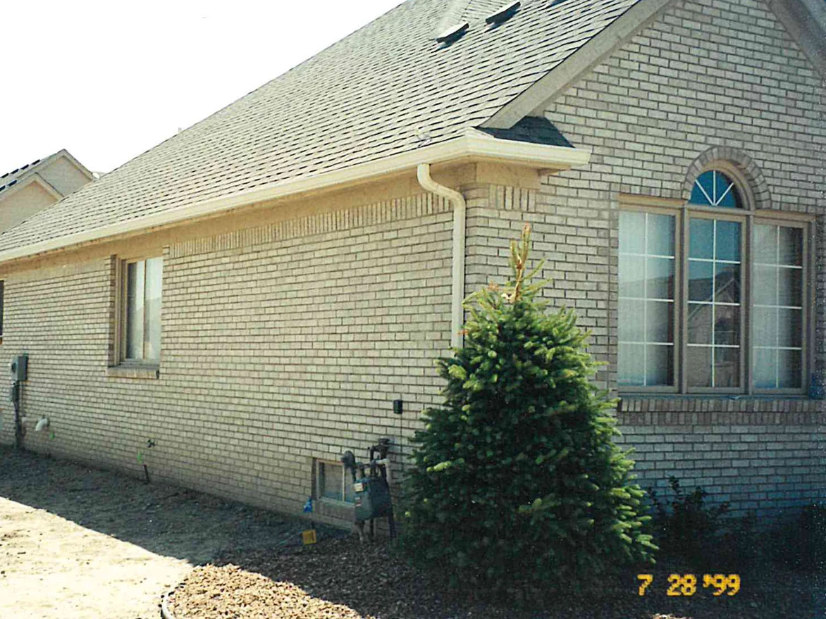The side elevation in 1999, a long run of cream brick under a dentil course, with a young spruce at the gable end and the ground still bare