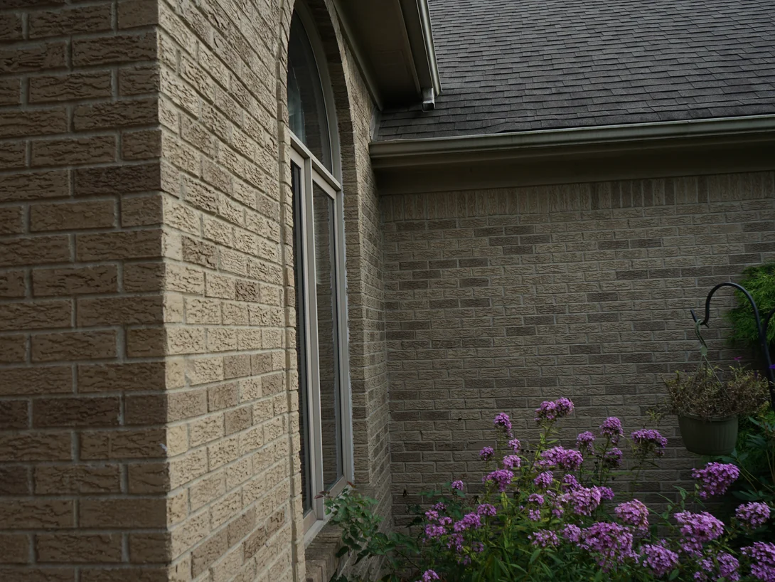 An outside corner of the house in 2019 with a round-headed window beyond, the brick an even tan range above a bed of phlox