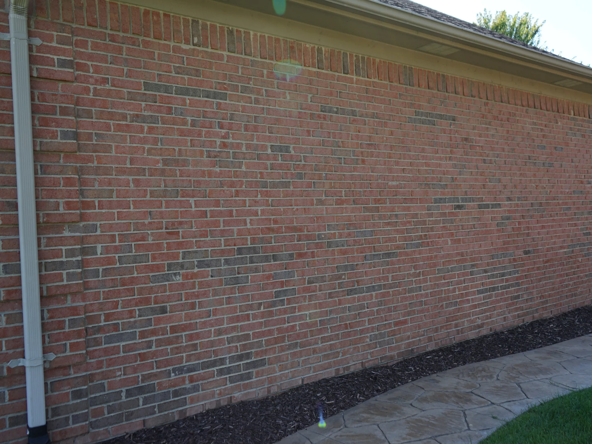 A long wall of the house in 2019 under a low hipped roof, the brick one even red range beside a stamped concrete walkway