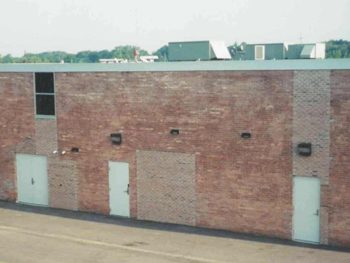 The long brick wall in 2000 before staining, with a bricked-in opening in the middle, a paler patch below the high window, a paler pier at the far end and three pale steel doors