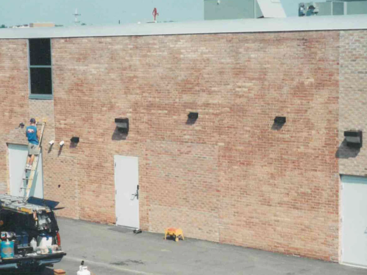 An applicator on an extension ladder working the brick beside the double door in 2000, a van with a roof rack and a drum on a tarp in the drive below him
