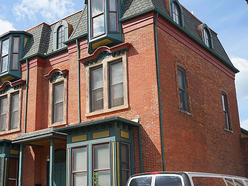 The front and the side of the home in 2019, one range of red brick from the porch bays up past the window hoods to the dormers, the woodwork in green and red
