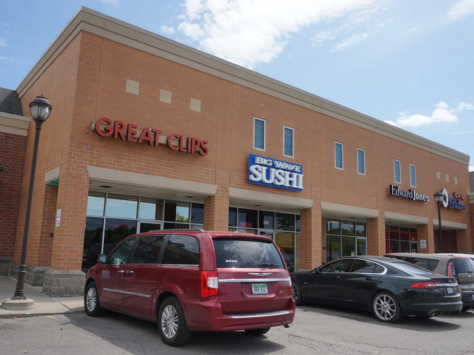 The plaza from the car park in 2019, lit tenant signs across the storefronts and the upper wall above them holding the same tan and red range as the piers at ground level.