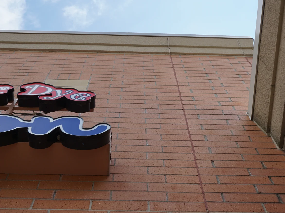 The upper wall in 2019 looked at from directly below, the courses running unbroken past a control joint to the coping, with the edge of a shopfront sign at the left