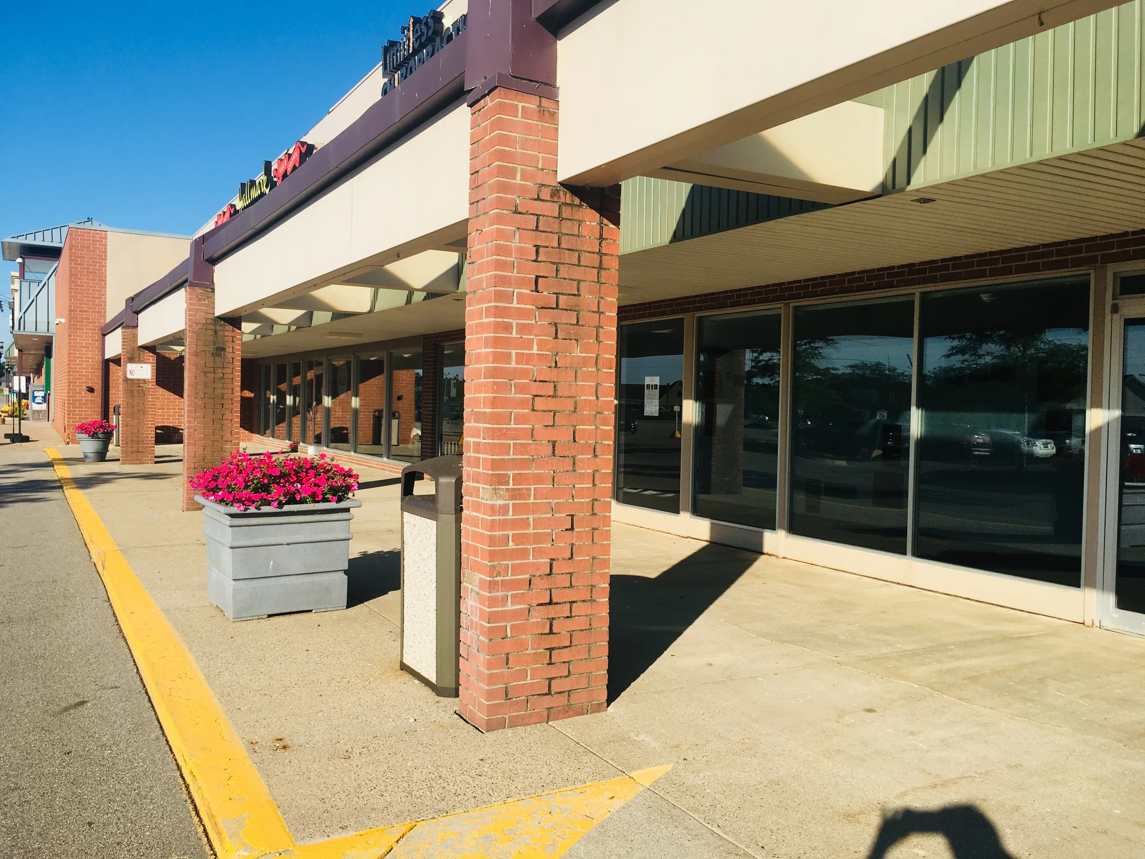 The plaza in 2019 seen along the walkway, brick piers stepping away past planters and shopfront glass, every pier holding the same red range