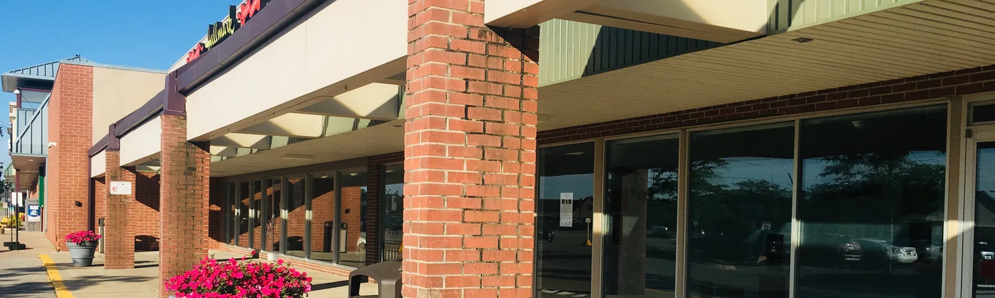 The shopfronts of a single-story plaza in 2019, its brick piers and low walls holding one red range along the walkway