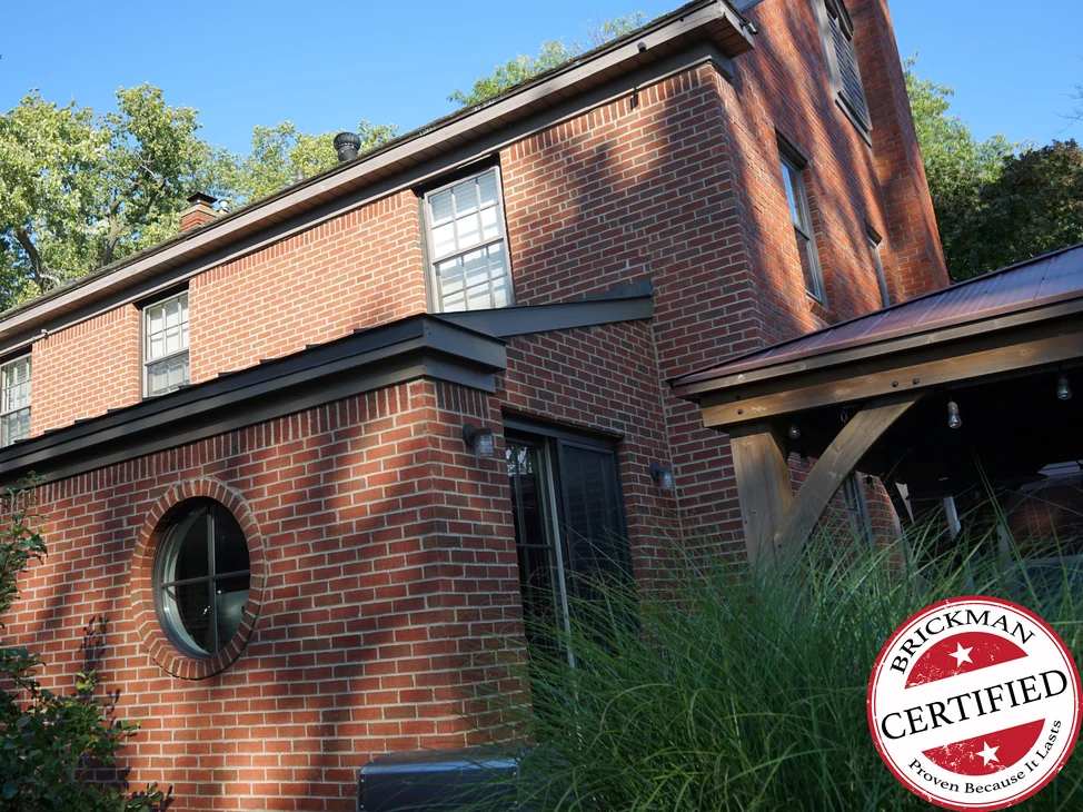 The house from the garden in summer sun in 2019, its brick an even red across every face, with a round window in a radial brick surround and a timber covered porch to the right.