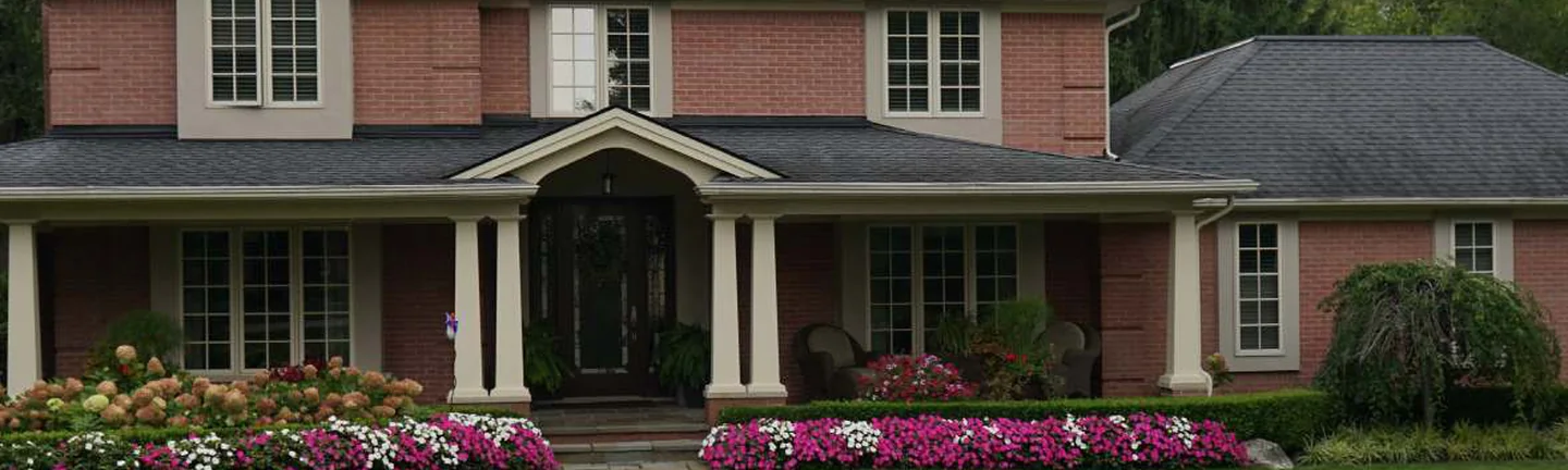 The front of the Michigan house in 2019, salmon brick between cream trim and columns, with beds of pink and white flowers along the porch