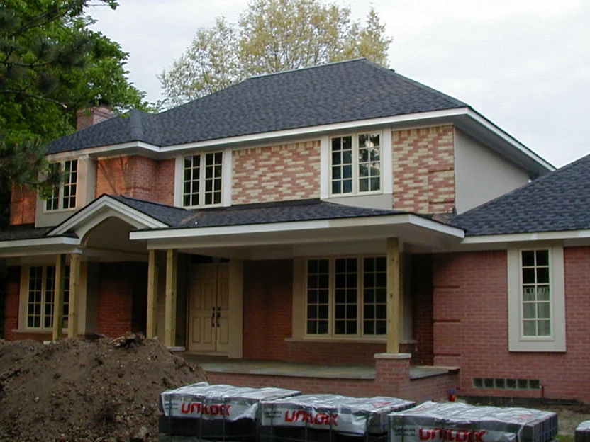 The house part way through staining in 2003, the upper storey a patchwork of yellow and salmon units around the windows while the lower storey is already one color, with a mound of earth and pallets of pavers on the drive