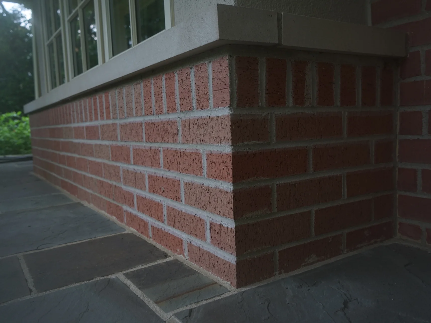 The corner of a stained brick base under a window on the porch, a cast stone cap on top and bluestone paving at its foot, shot from low on the porch floor
