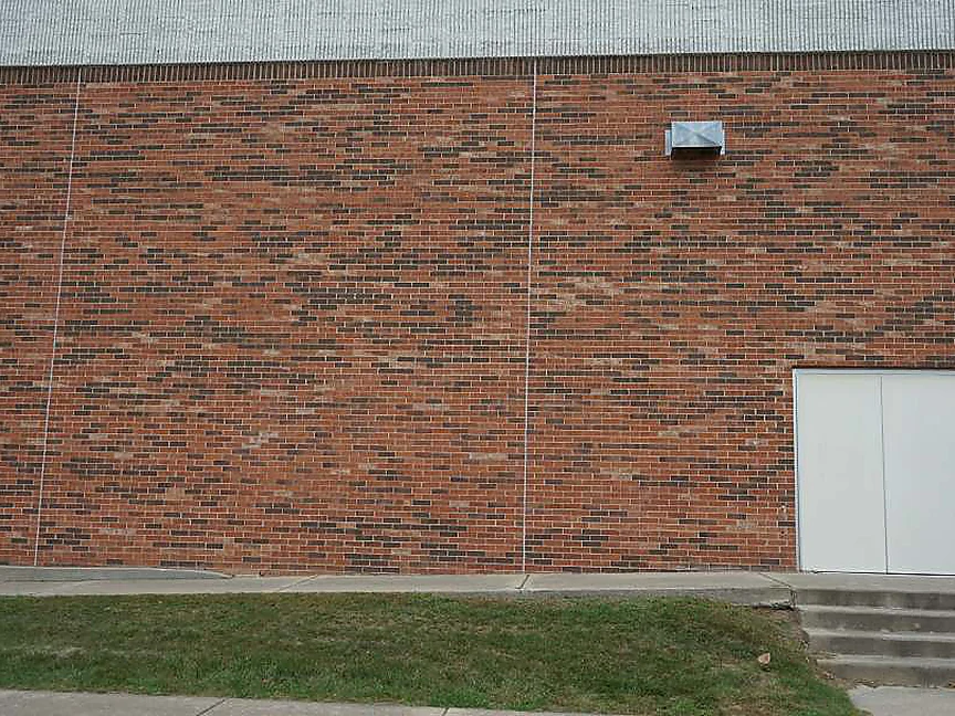 A flat elevation of the school in 2019, red brick with dark headers through every course, two vertical control joints, a metal vent on the face and a white double service door with concrete steps at the right