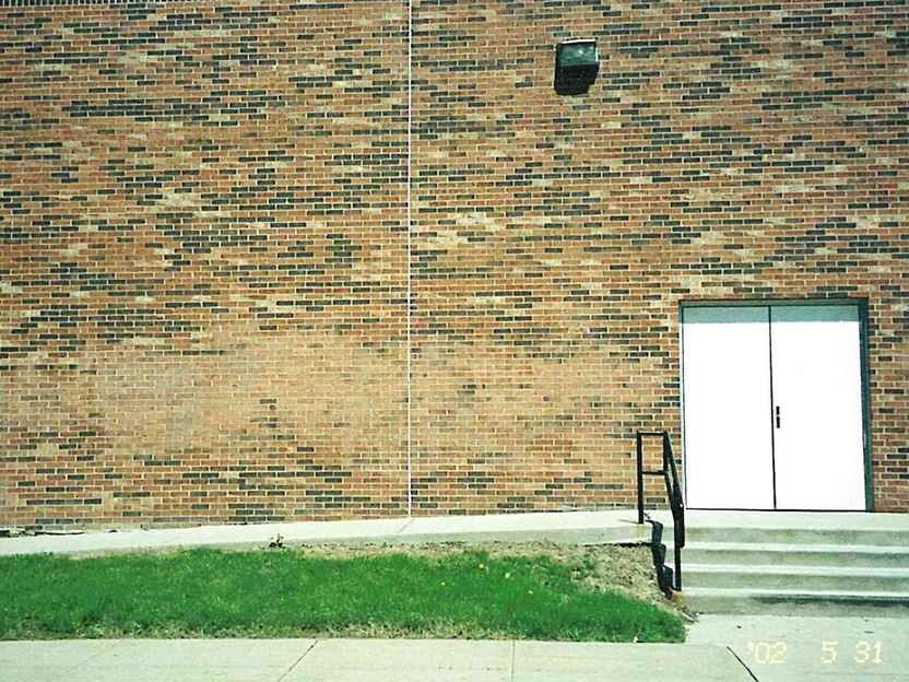 A blank elevation of the school in 2002, tan and salmon brick with darker units scattered unevenly through the courses, a single wall light, a white double service door at the right and a grass strip along the walk