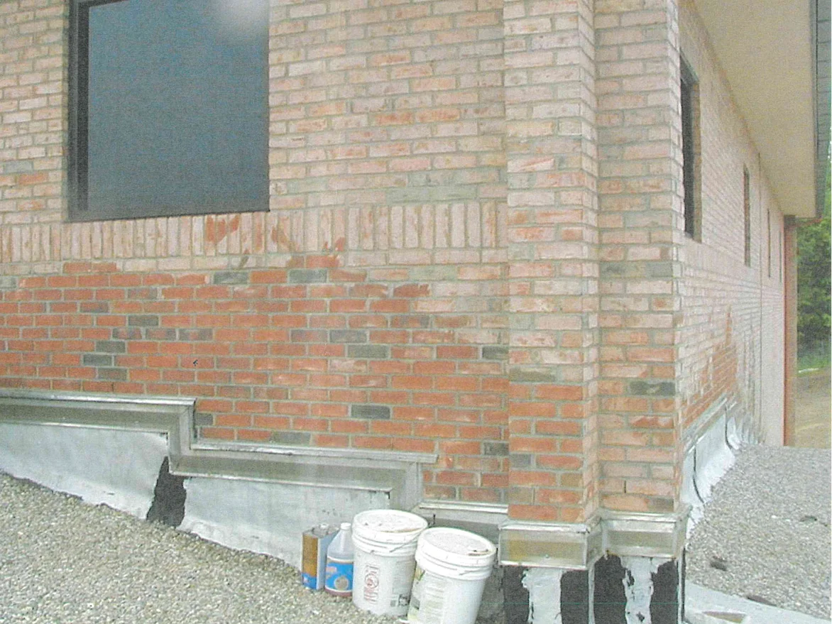 An outside corner of the school before the work, the cream brick above patchy and washed out and the courses below it a raw orange red, with buckets standing on the gravel