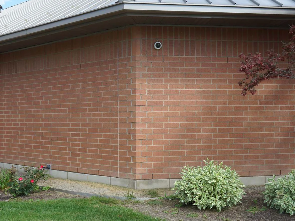 An outside corner of the Michigan building in 2019, red brick running through the shaped corner units and on across both walls under a standing seam metal roof, with mown lawn and shrubs along the foot