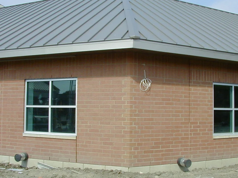 The outside corner of the single story building in 2002, the shaped units running up the corner reading pink and flat against the red brick of both walls, bare dirt at the base and a coil of white wire hung beside the corner
