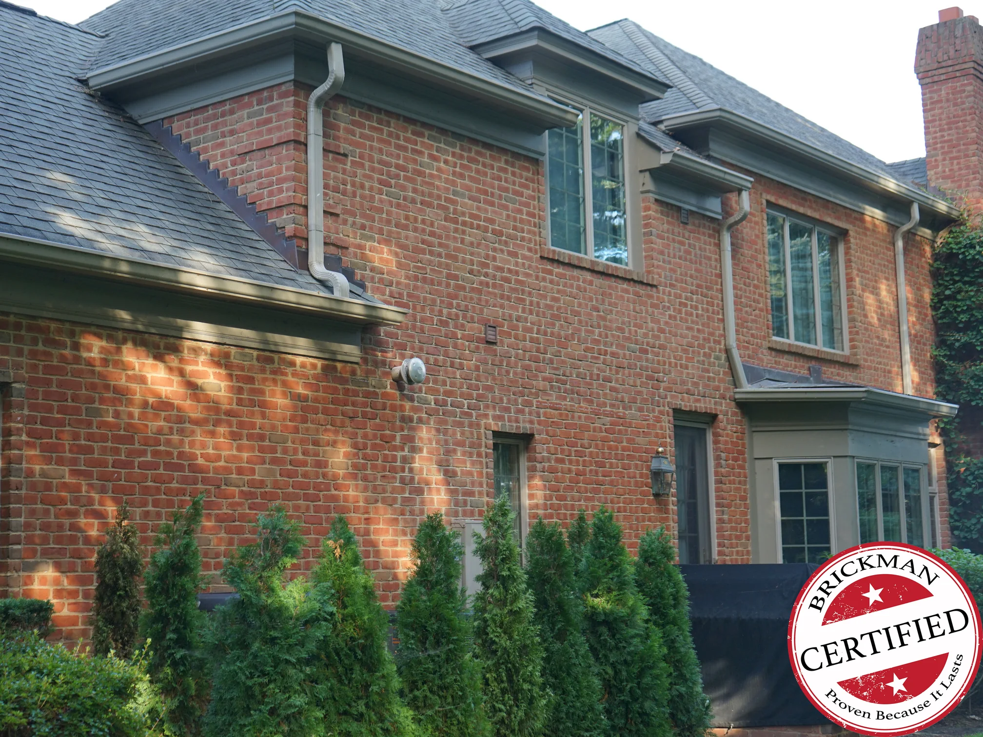 The side of the house in 2019, evening sun across an even red brown brick range, the brickwork stepping down the roof slope at the left and a Brickman Certified stamp printed over the lower right of the frame