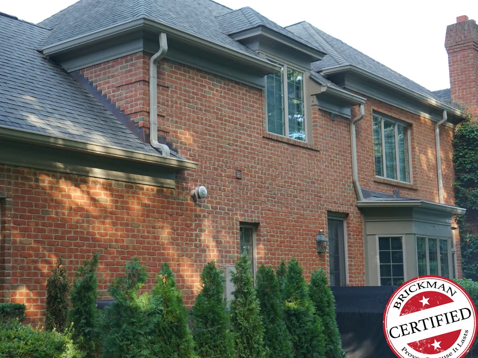 The side of the house in 2019, evening sun across an even red brown brick range, the brickwork stepping down the roof slope at the left and a Brickman Certified stamp printed over the lower right of the frame.