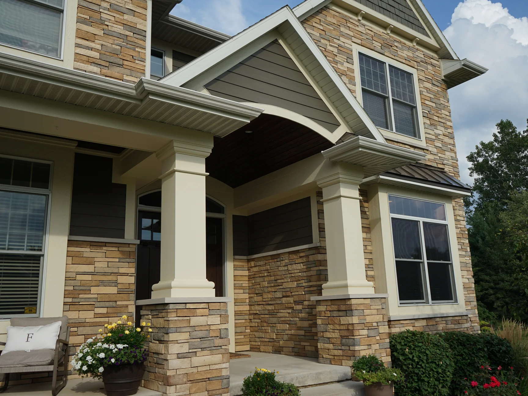 Looking up at the front entry in 2019, two cream columns on stone piers under a gabled porch roof, the stone veneer carrying up to the second floor windows in one tan and grey blend
