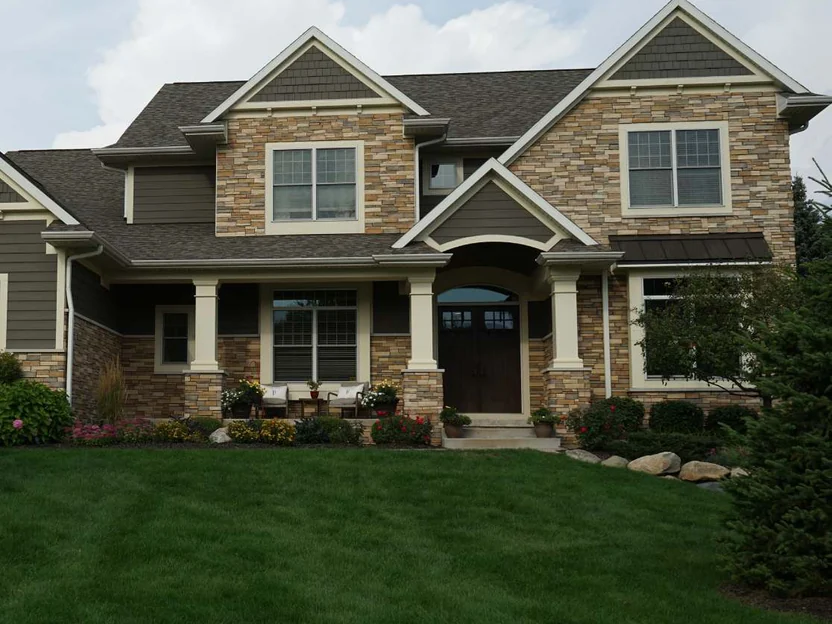 The whole front of the two-story house in 2019, stone veneer on both gables and the porch piers, dark siding above, a lawn and planted beds in front