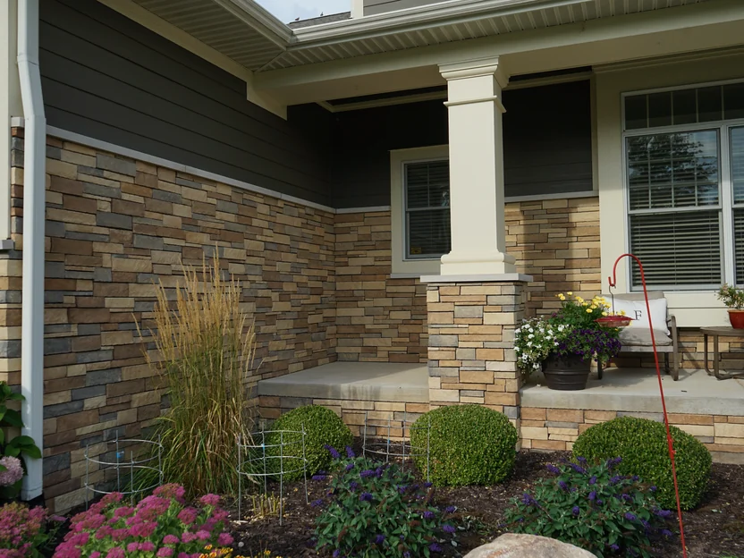 The porch corner in 2019, the same wall that held the sample boards in 2012, with a column on a stone pier, a bench, potted flowers and clipped shrubs in front