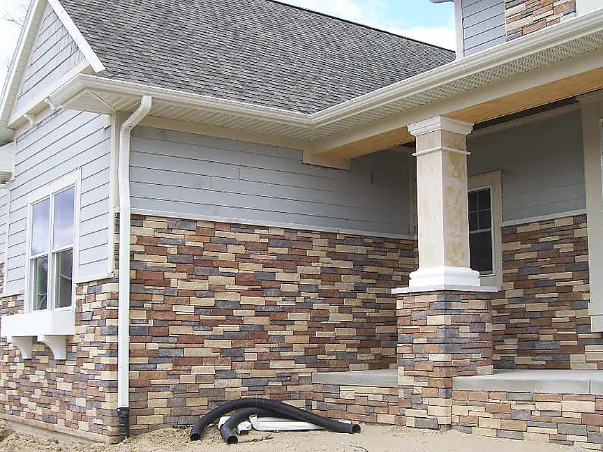 The side wall of the house in 2012 before staining, grey siding over a stone veneer base of separate rust, charcoal and cream units, a porch column at the corner and drain pipe on the sand