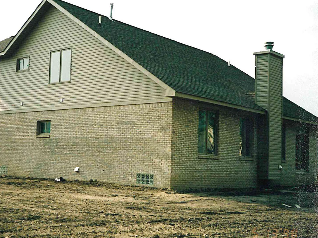 The side of the house in 1998 with the yard still bare earth, light tan brick to the first floor and siding in the gable above, the roof sweeping down to the right over a single-story wing