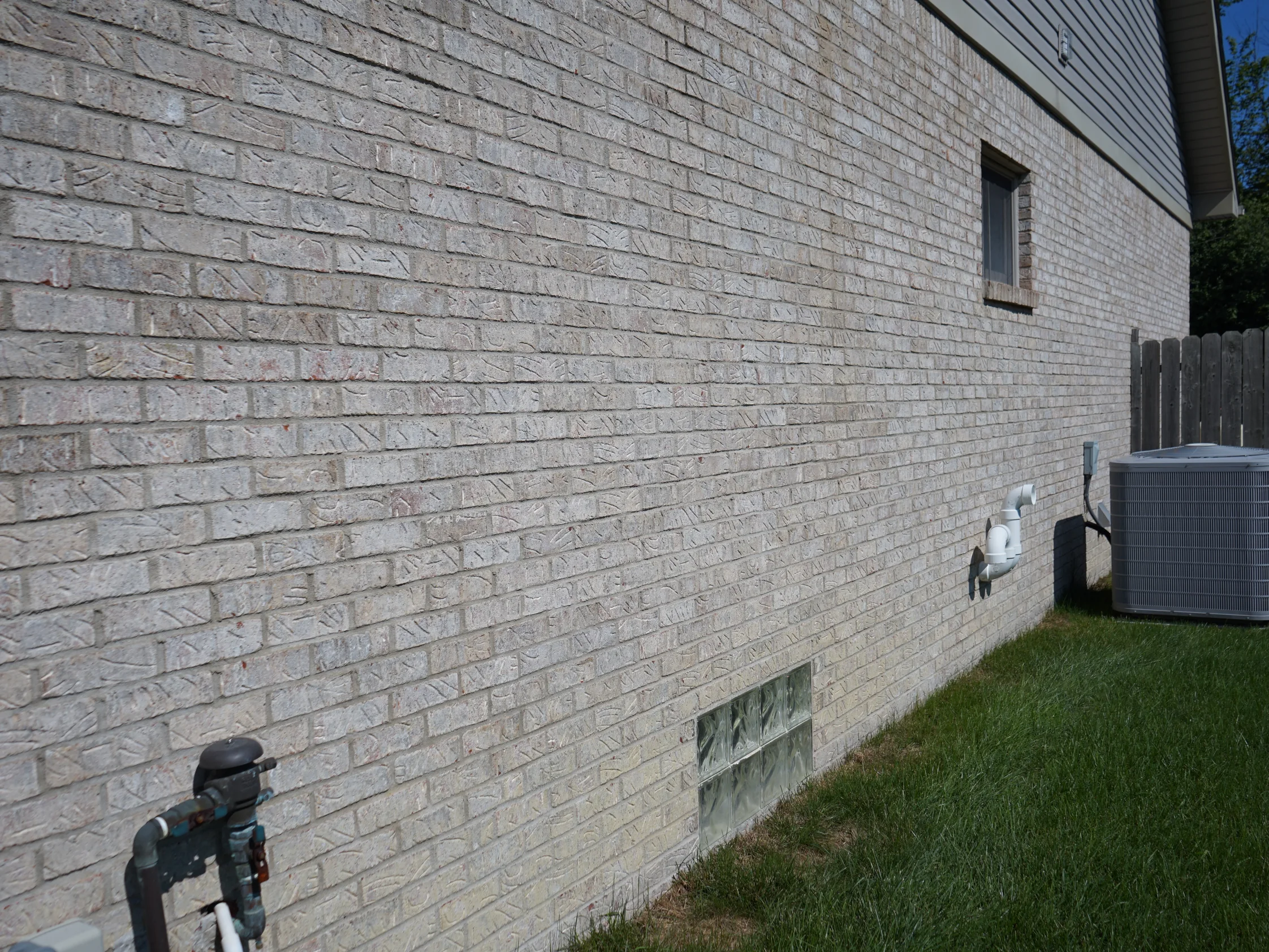 The side wall from the rear corner in 2019, the brick running unbroken past a basement window and a glass block light to the far end