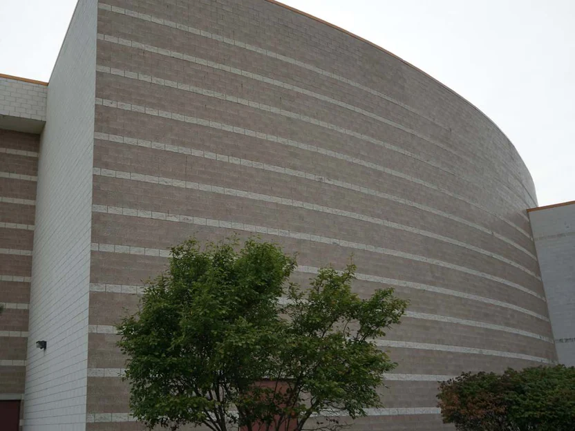 The curved wall from its outside corner in 2019, tan block banded with pale accent courses, a white block wing and a red door at the left and a small tree in front